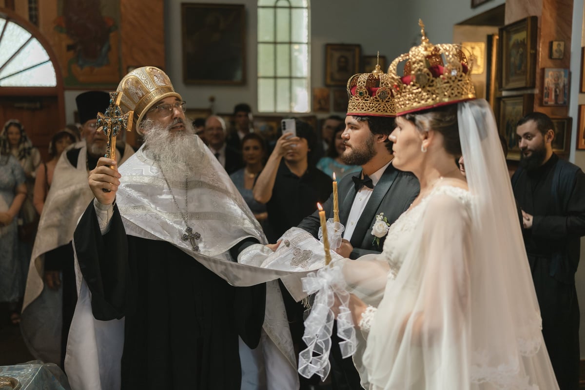 A crowned Orthodox bride and groom follow a priest during the wedding procession