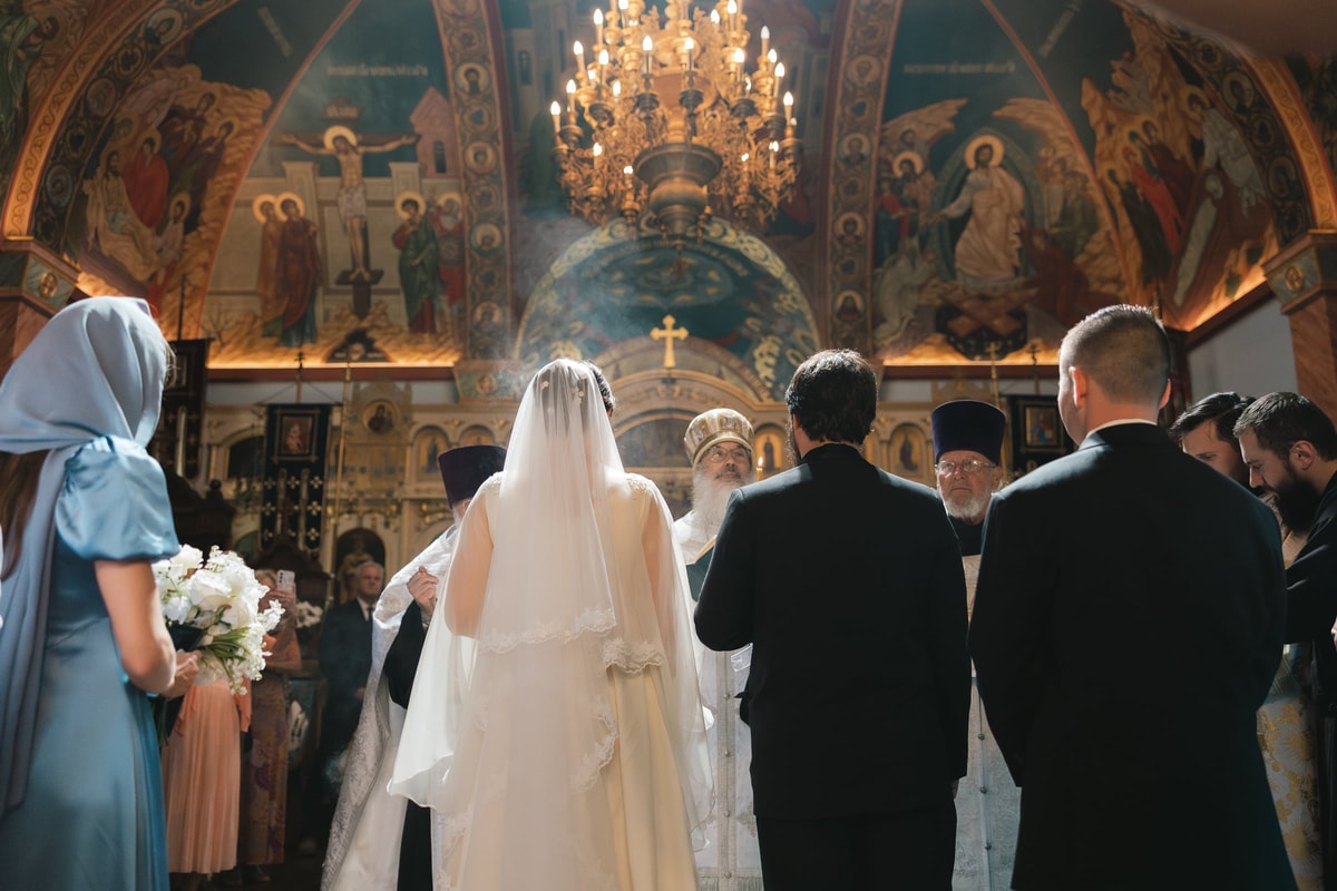 A bride and groom stand before the iconostasis with Orthodox clergy