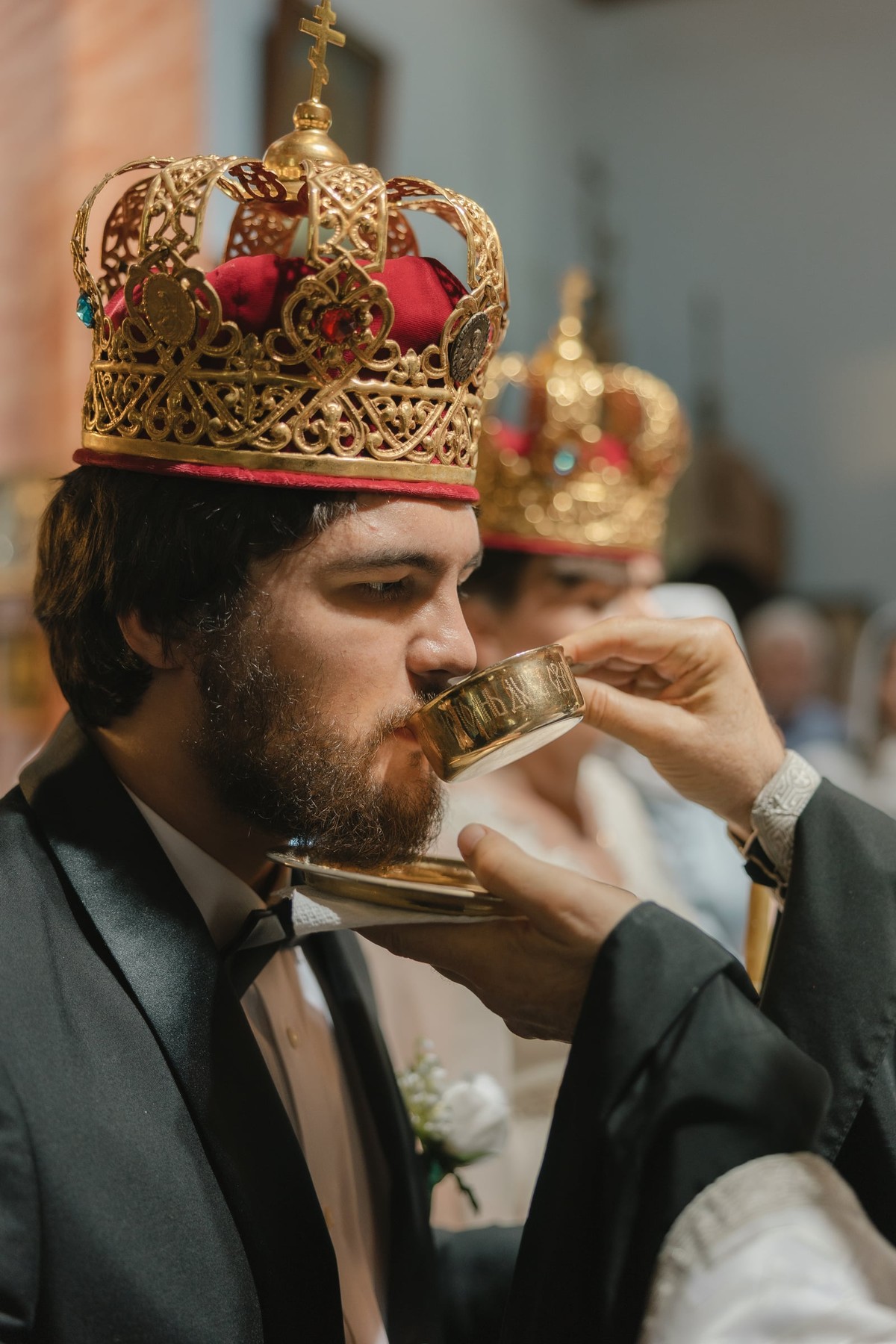 The groom receives the common cup while wearing his wedding crown