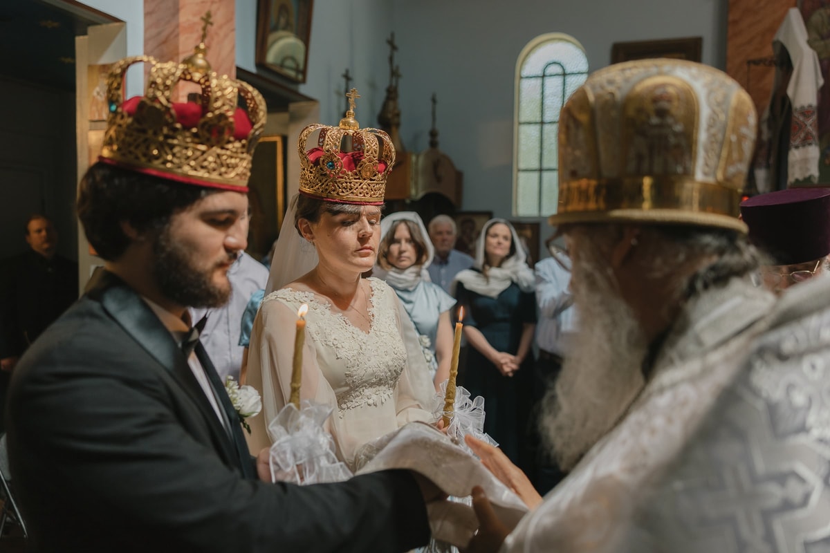 The bride and groom listen to the bishop while wearing their crowns