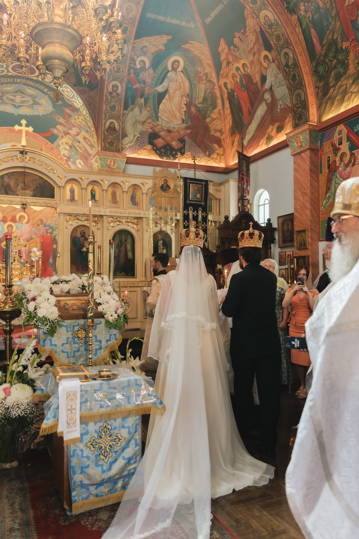 A bride and groom stand before Orthodox clergy beneath painted church murals