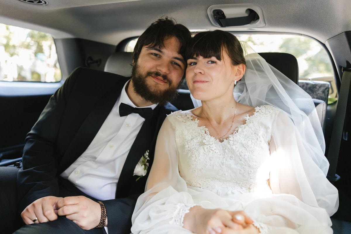 The bride and groom smile together in the back seat of a car