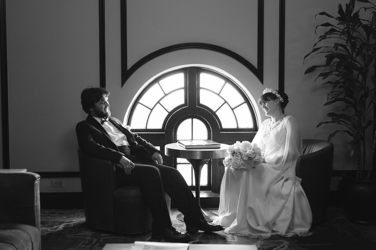 The bride and groom sit together before a round window in a black-and-white portrait