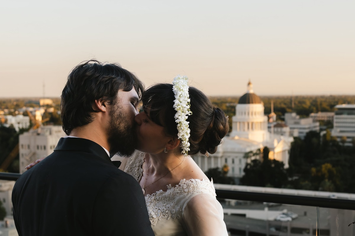 A bride and groom kiss with a domed capitol building behind them