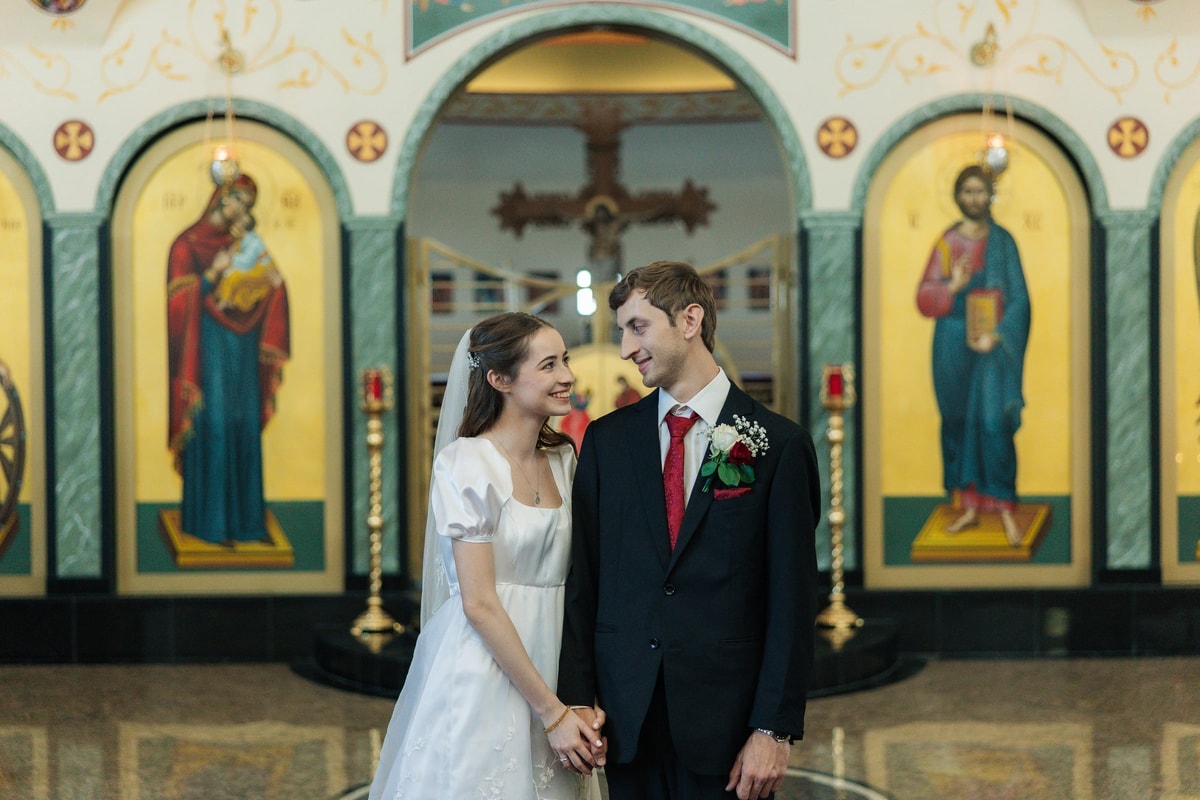 The bride and groom smile at one another before the iconostasis