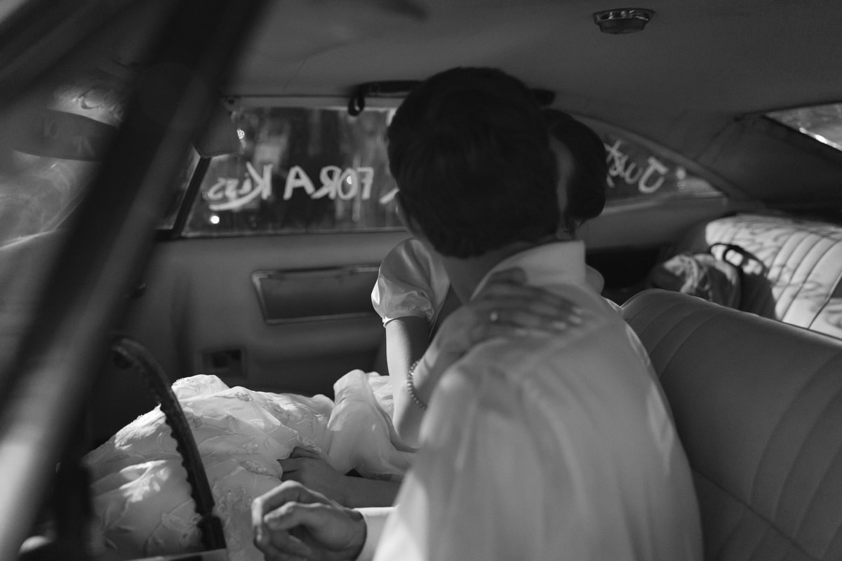 The bride and groom kiss inside the getaway car in a black-and-white photograph