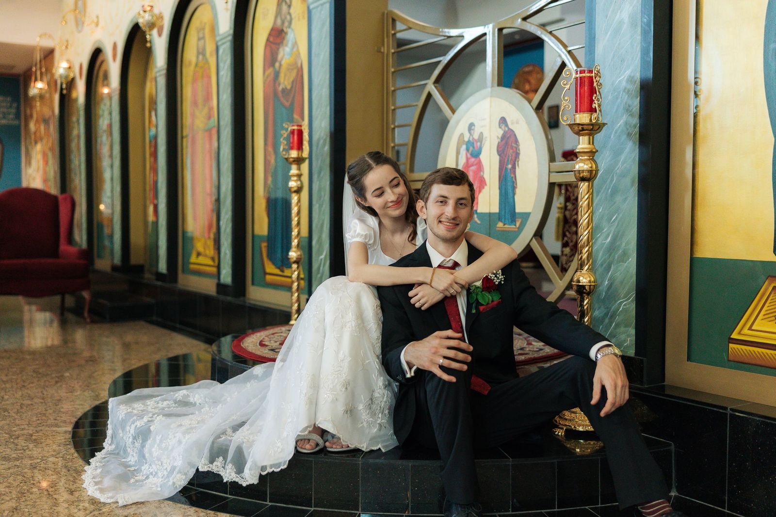 A bride sits behind the groom with her arms around his shoulders inside an Orthodox church